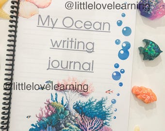 May include: A spiral-bound notebook labelled "My Ocean writing journal" featuring a vibrant coral reef illustration. The image includes ocean-themed decorations: a starfish, seashells, and fish-shaped ornaments, arranged around the journal.