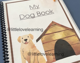 May include: A spiral-bound book titled "My Dog Book" with a watercolour illustration of a golden retriever and dog house. A small, orange-brown plastic dog figurine sits on top of the book. The book has a blue cover.