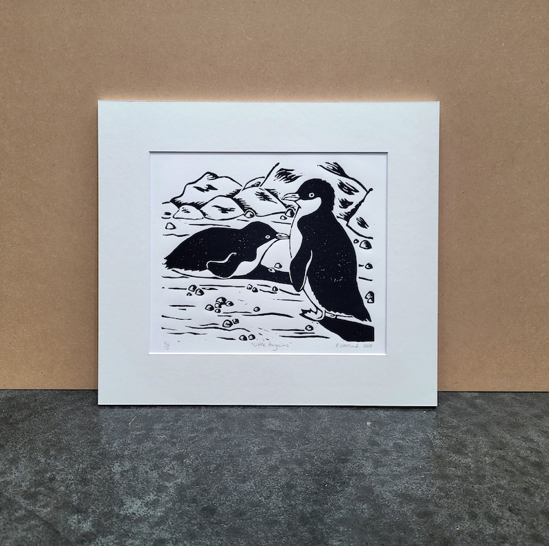 Puede incluir: Grabado en lin&oacute;leo en blanco y negro de dos ping&uuml;inos en una costa rocosa. La impresi&oacute;n est&aacute; titulada "Peque&ntilde;os ping&uuml;inos" y est&aacute; firmada y fechada "F. Smith 2023".