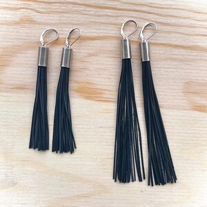 May include: Two pairs of black tassel earrings. Each earring features a silver-colored metal top and a cascade of thin, black strands. The earrings are displayed on a light-colored wooden surface.
