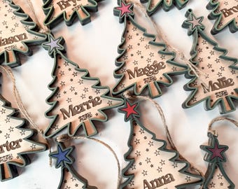 May include: Wooden Christmas tree ornaments with names and the year 2023 engraved. Each ornament has a star on top and a star pattern. The ornaments are light brown with green edges and are strung with twine.