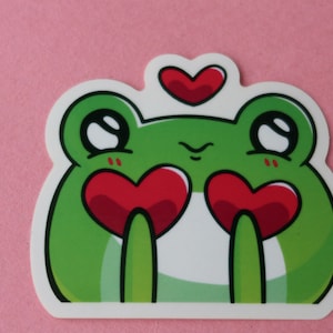 Puede incluir: Una pegatina de rana verde con ojos grandes y un pequeño corazón rojo sobre su cabeza. La rana sostiene dos corazones rojos grandes en sus manos.