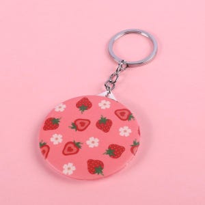 May include: A pink round keychain with a silver key ring. The keychain has a white background with a repeating pattern of red strawberries and white flowers.