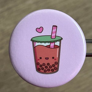 May include: A purple button with a cartoon image of a boba tea with a pink and white striped straw and a green lid. The boba tea has a smiling face and brown boba pearls. A small pink heart is above the boba tea.