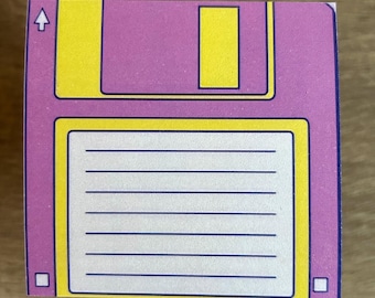 May include: A pink and yellow floppy disk shaped sticky note pad. The pad has a blue outline and a white lined area for writing.