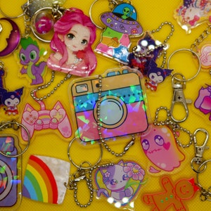 May include: A collection of colourful, holographic keychains featuring various cartoon characters, including a pink and blue camera, a rainbow, a gingerbread person, a ghost, a cat, and a unicorn.