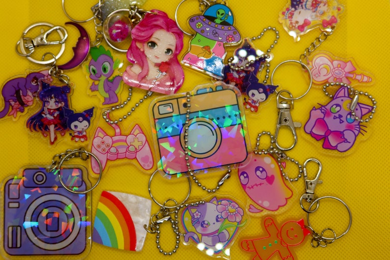 May include: A collection of colourful, holographic keychains featuring various cartoon characters, including a pink and blue camera, a rainbow, a gingerbread person, a ghost, a cat, and a unicorn.