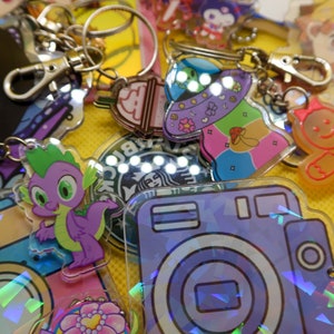 May include: A collection of colourful keychains featuring various characters and designs, including a purple dragon, a gingerbread person, a camera, and a Starbucks logo. The keychains are made of clear acrylic and have a holographic finish.