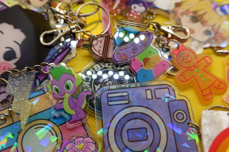 May include: A collection of colourful keychains featuring various characters and designs, including a purple dragon, a gingerbread person, a camera, and a Starbucks logo. The keychains are made of clear acrylic and have a holographic finish.
