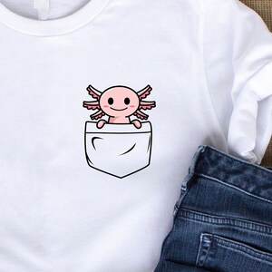 Puede incluir: Camiseta blanca con un bolsillo que presenta un axolotl de dibujos animados. El axolotl es rosa con ojos negros y una peque&ntilde;a sonrisa.