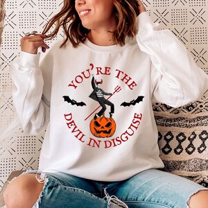 May include: White sweatshirt with a red graphic print of a devil standing on a pumpkin with the text "You're the Devil in Disguise".