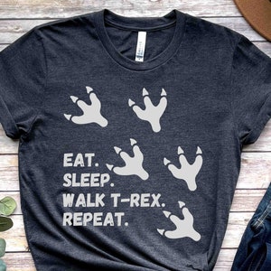 May include: A dark gray t-shirt with a white graphic of dinosaur footprints and the text "Eat. Sleep. Walk T-Rex. Repeat."