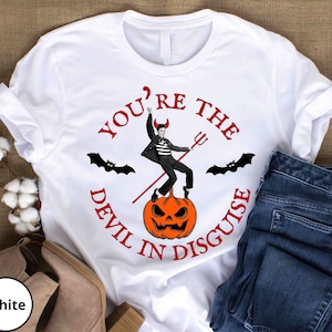May include: White t-shirt with a red, black, and orange graphic. The graphic features a man in a black and white costume standing on a jack-o-lantern. The text "You're the Devil in Disguise" is written around the graphic.