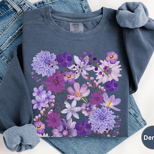 May include: A dark blue sweatshirt with a floral print of purple and white flowers. The flowers are arranged in a bouquet shape and are centered on the front of the sweatshirt.