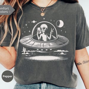 May include: Dark grey t-shirt featuring a white graphic of an alien in a UFO, holding a phone and giving a peace sign. The design includes a desert landscape, stars, and a planet. The shirt is a comfort color.