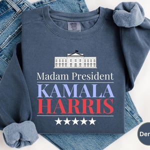 May include: A navy blue sweatshirt with the text "Madam President Kamala Harris" and a white silhouette of the White House. The text is in red, white, and blue.