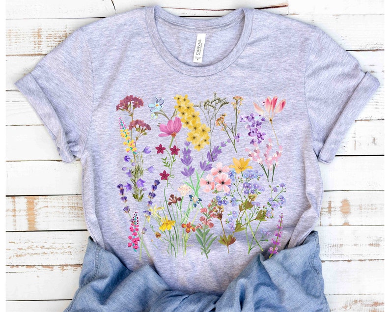 Może przedstawiać: Szary t-shirt z kolorowym nadrukiem kwiatowym przedstawiającym r&oacute;żne kwiaty w odcieniach r&oacute;żowego, fioletowego, ż&oacute;łtego i niebieskiego.