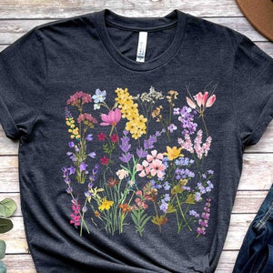 Może przedstawiać: Szary t-shirt z kolorowym nadrukiem kwiatowym z r&oacute;żowymi, fioletowymi, ż&oacute;łtymi i niebieskimi kwiatami. Kwiaty są ułożone w skupisko i otoczone zielonymi liśćmi.