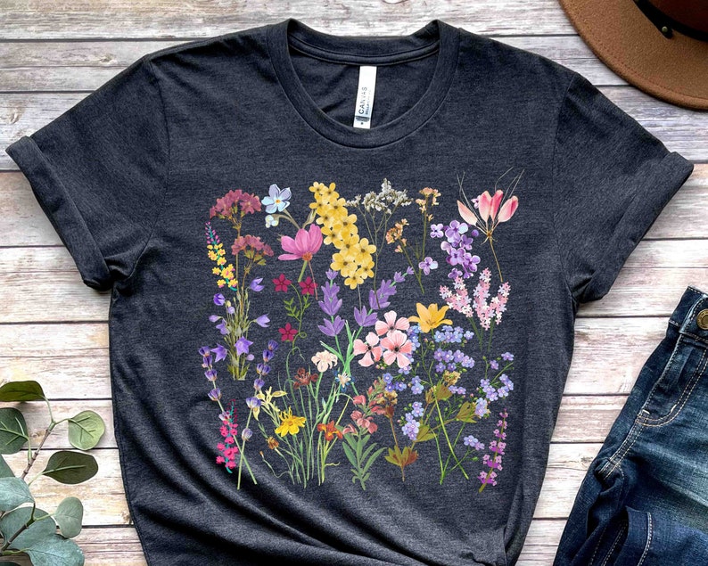 Może przedstawiać: Szary t-shirt z kolorowym nadrukiem kwiatowym z r&oacute;żowymi, fioletowymi, ż&oacute;łtymi i niebieskimi kwiatami. Kwiaty są ułożone w skupisko i otoczone zielonymi liśćmi.