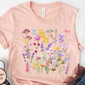 Może przedstawiać: Jasnor&oacute;żowy t-shirt z nadrukiem kwiatowym przedstawiającym r&oacute;żne kwiaty w odcieniach fioletu, r&oacute;żu, ż&oacute;łtego i niebieskiego. Kwiaty są ułożone w skupisko na przodzie koszulki.