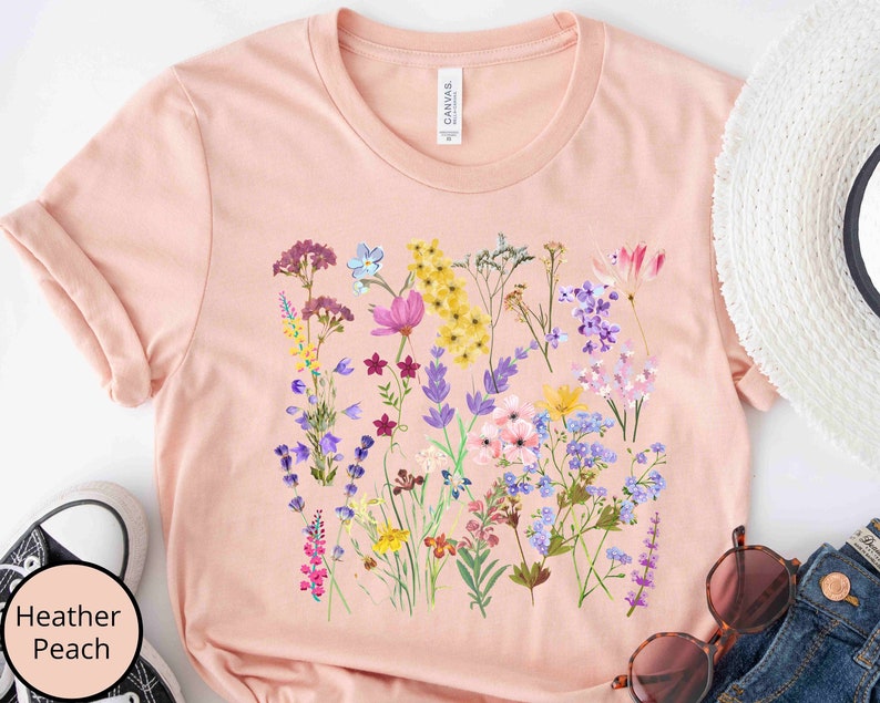 Może przedstawiać: Jasnor&oacute;żowy t-shirt z nadrukiem kwiatowym przedstawiającym r&oacute;żne kwiaty w odcieniach fioletu, r&oacute;żu, ż&oacute;łtego i niebieskiego. Kwiaty są ułożone w skupisko na przodzie koszulki.