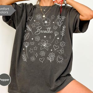 May include: A dark gray oversized t-shirt with a white floral design and the word "Breathe" in the center. The shirt has a relaxed fit and a crew neck.