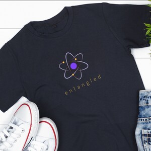 May include: Dark navy blue t-shirt featuring a graphic of an atom with a purple center and yellow accents. The word "entangled" is printed in gold below the atom design. White canvas sneakers with red trim are visible.