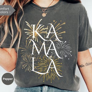 May include: A gray t-shirt with a white graphic of the word "KAMALA" surrounded by gold glitter fireworks.