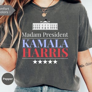 May include: A dark gray t-shirt with a white silhouette of the White House and the text "Madam President Kamala Harris" in blue, red, and white lettering. Five white stars are below the text.