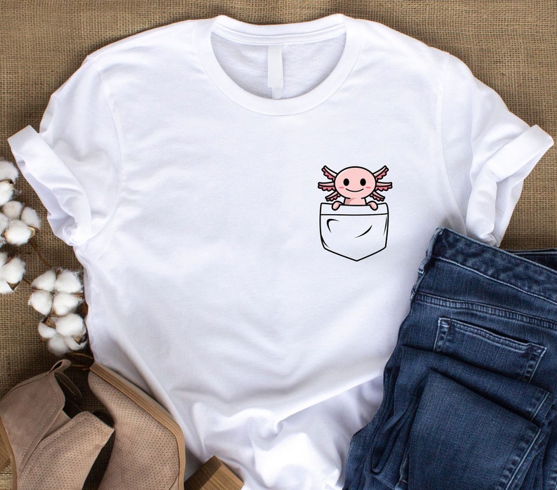 Puede incluir: Camiseta blanca con un bolsillo que presenta un axolotl de dibujos animados. El axolotl es rosa con ojos negros y una peque&ntilde;a sonrisa.