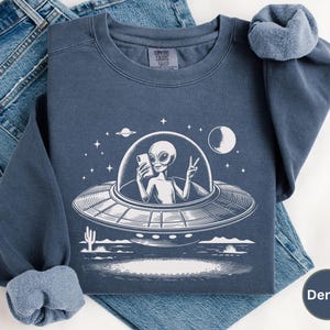 May include: A dark blue sweatshirt with a white graphic of an alien in a flying saucer holding a phone and making a peace sign. The alien is wearing a spacesuit and has a large head. The graphic is surrounded by stars and planets. The sweatshirt is made of a soft, comfortable material and is perfect for everyday wear.