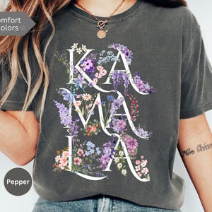 May include: Dark gray t-shirt with the word "KAMALA" in white, filled with colorful floral arrangements. The shirt has a crew neck and short sleeves. The text "Comfort Colors" and the color "Pepper" are visible.