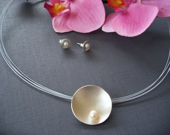 May include: A silver necklace with a delicate chain and a round pendant featuring a pearl. The pendant is a smooth, curved disc with a subtle shine. The necklace is accompanied by a pair of matching silver earrings with small pearls.
