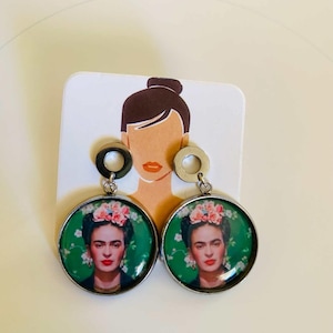 Könnte beinhalten: Ein Paar runde, silberfarbene Ohrhänger mit einem Porträt von Frida Kahlo mit Blumenschmuck vor grünem Hintergrund. Die Ohrringe sind auf einer weißen Karte mit einer stilisierten Illustration eines Kopfes befestigt.