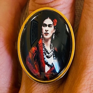 Può includere: Un anello di forma ovale con un ritratto lucido e ad alto contrasto di Frida Kahlo. L'immagine mostra una persona con un fiore rosso tra i capelli, che indossa uno scialle rosso scuro e collane di perline a strati su sfondo nero, incastonato in una cornice di metallo dorato.