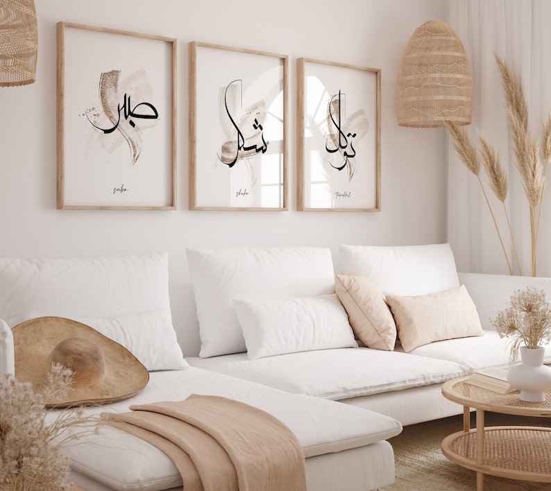 May include: Three framed prints with Arabic calligraphy in black ink on a white background. The prints are hung on a white wall above a white sectional sofa with beige throw pillows and a beige throw blanket.