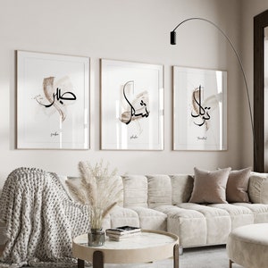 May include: Three framed prints with Arabic calligraphy in black ink on a white background. The prints are hung on a white wall above a white couch with a brown throw blanket and a vase of dried pampas grass.
