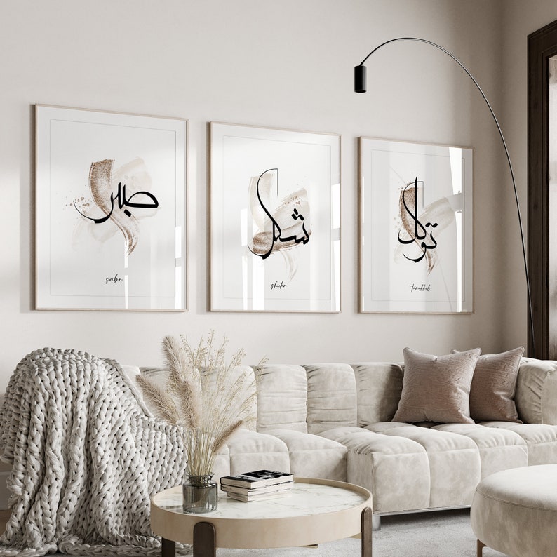 May include: Three framed prints with Arabic calligraphy in black ink on a white background. The prints are hung on a white wall above a white couch with a brown throw blanket and a vase of dried pampas grass.