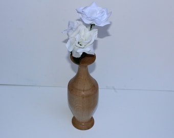 May include: A wooden vase with a light brown finish, holding two white roses. The vase has a bulbous body and a flared base, with a narrower neck and a circular top. The roses are in full bloom, adding a touch of elegance.