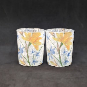 May include: Two clear glass candle holders with a white background and a floral pattern. The pattern features yellow flowers and blue flowers.