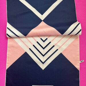 May include: A rectangular Nina Ricci Paris scarf featuring a geometric pattern of navy blue, soft pink, and cream white stripes and triangles. The scarf has a smooth texture, a fringed bottom edge, and a white label with black text at the base.