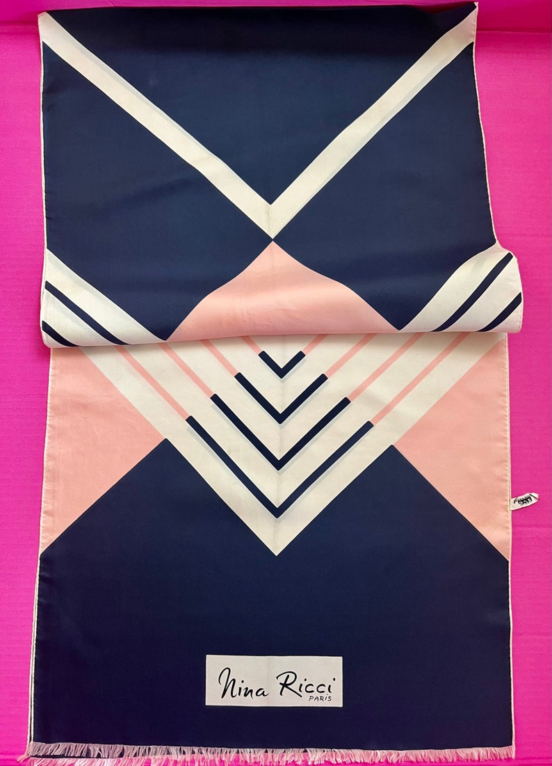 May include: A rectangular Nina Ricci Paris scarf featuring a geometric pattern of navy blue, soft pink, and cream white stripes and triangles. The scarf has a smooth texture, a fringed bottom edge, and a white label with black text at the base.