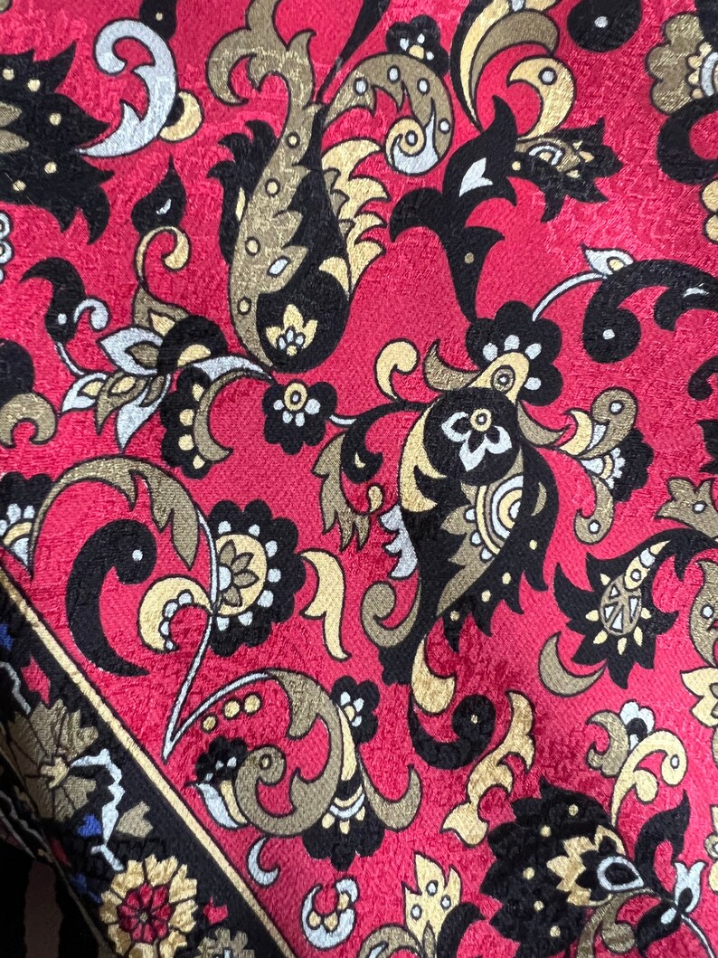 Puede incluir: Una tela con un fondo rojo vibrante y un patr&oacute;n repetitivo intrincado de motivos de cachemira y elementos florales estilizados en negro, dorado y blanco. El dise&ntilde;o incluye enredaderas y formas detalladas en la superficie.
