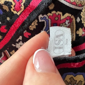 Puede incluir: Una prenda de tela negra con un patr&oacute;n vibrante e intrincado de estilo cachemir en tonos rojo, azul y dorado. Una etiqueta de ropa rectangular blanca est&aacute; unida a la tela, mostrando el texto "ISA" en una fuente sans-serif en negrita.
