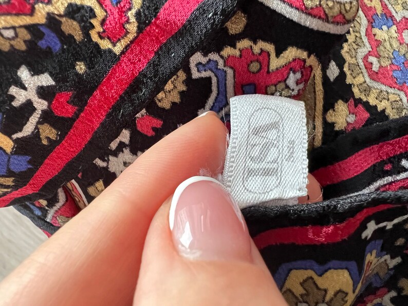 Puede incluir: Una prenda de tela negra con un patr&oacute;n vibrante e intrincado de estilo cachemir en tonos rojo, azul y dorado. Una etiqueta de ropa rectangular blanca est&aacute; unida a la tela, mostrando el texto "ISA" en una fuente sans-serif en negrita.