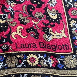 Puede incluir: Un tejido con un fondo rojo vibrante y un borde negro, decorado con intrincados patrones florales y de cachemira en oro, negro y azul. El texto Laura Biagiotti est&aacute; impreso en letras negras sobre la secci&oacute;n roja de la tela.