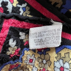 Puede incluir: Una tela de seda estampada con un dise&ntilde;o floral en negro, rojo, azul y dorado. Una etiqueta de cuidado blanca est&aacute; adherida, mostrando el texto 100% SILK, s&iacute;mbolos de cuidado y MADE IN ITALY. La tela tiene una superficie texturizada y borde.