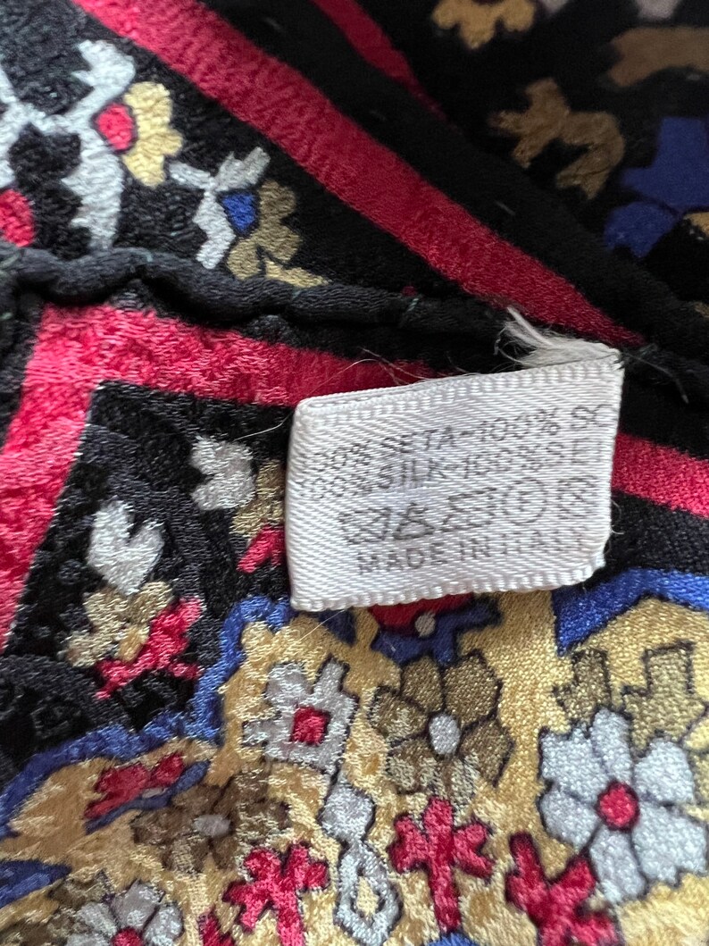 Puede incluir: Una tela de seda estampada con un dise&ntilde;o floral en negro, rojo, azul y dorado. Una etiqueta de cuidado blanca est&aacute; adherida, mostrando el texto 100% SILK, s&iacute;mbolos de cuidado y MADE IN ITALY. La tela tiene una superficie texturizada y borde.