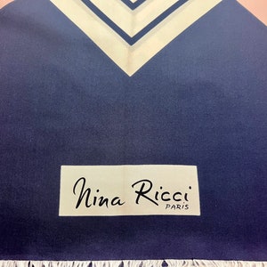 May include: A navy blue scarf featuring a white and navy chevron pattern at the top edge and a white rectangular label with the black text Nina Ricci PARIS printed on the lower section, finished with a white fringe along the bottom edge.