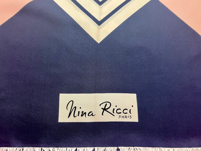 May include: A navy blue scarf featuring a white and navy chevron pattern at the top edge and a white rectangular label with the black text Nina Ricci PARIS printed on the lower section, finished with a white fringe along the bottom edge.